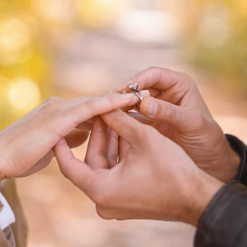 Man putting engagement ring on woman’s hand outdoors Close up of man putting engagement ring on woman's hand over blurred autumn forest background