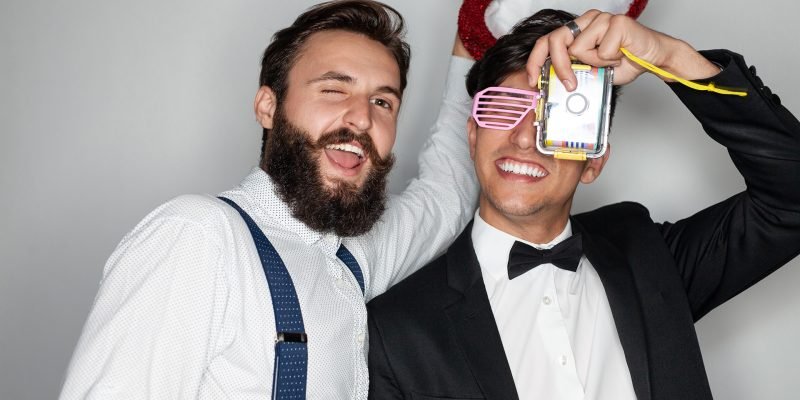 Happy young diverse guys having fun during party Content young multiracial male friends in stylish classy clothes smiling brightly and taking photo with film camera during party